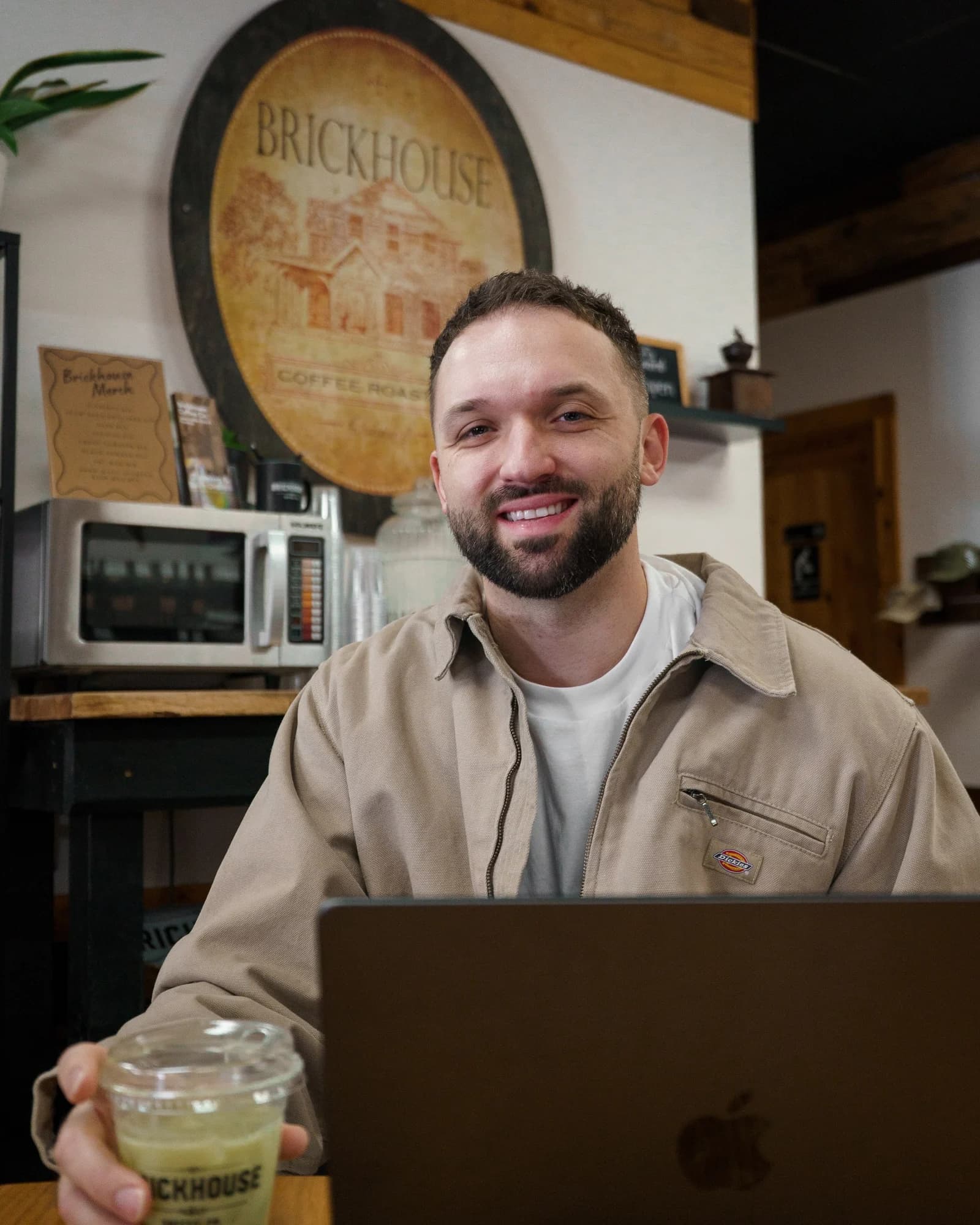 Kyle Hoffman smiling at a laptop in a coffee shop, holding an iced drink