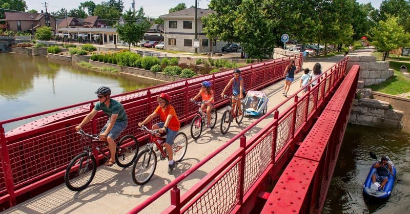Cyclists and a stroller crossing a red pedestrian bridge over the canal in Broad Ripple, with a kayaker below